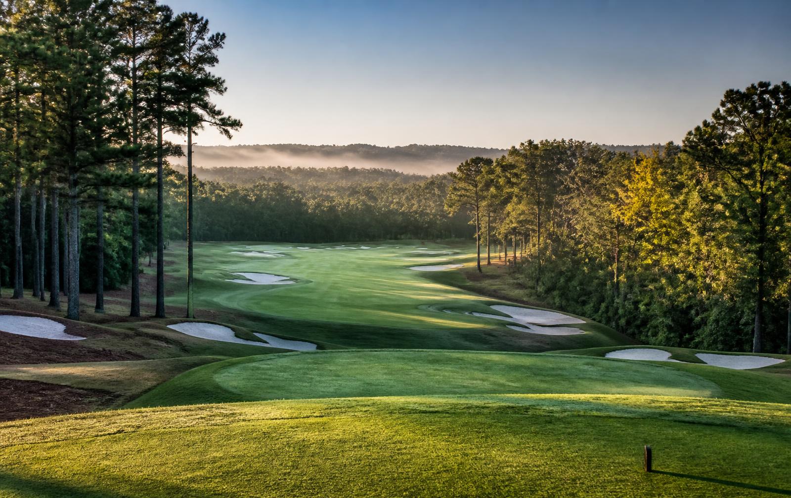 A championship fairway at sunrise seen from an elevated tee, framed by pines and white sand bunkers