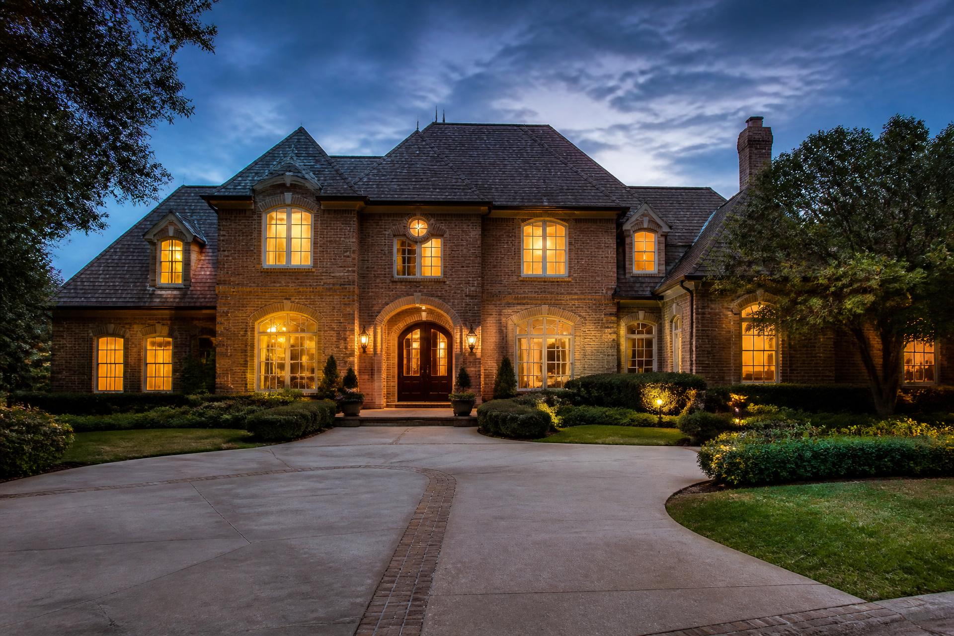 Front elevation of a large traditional brick luxury home with arched entry and mature landscaping at blue hour
