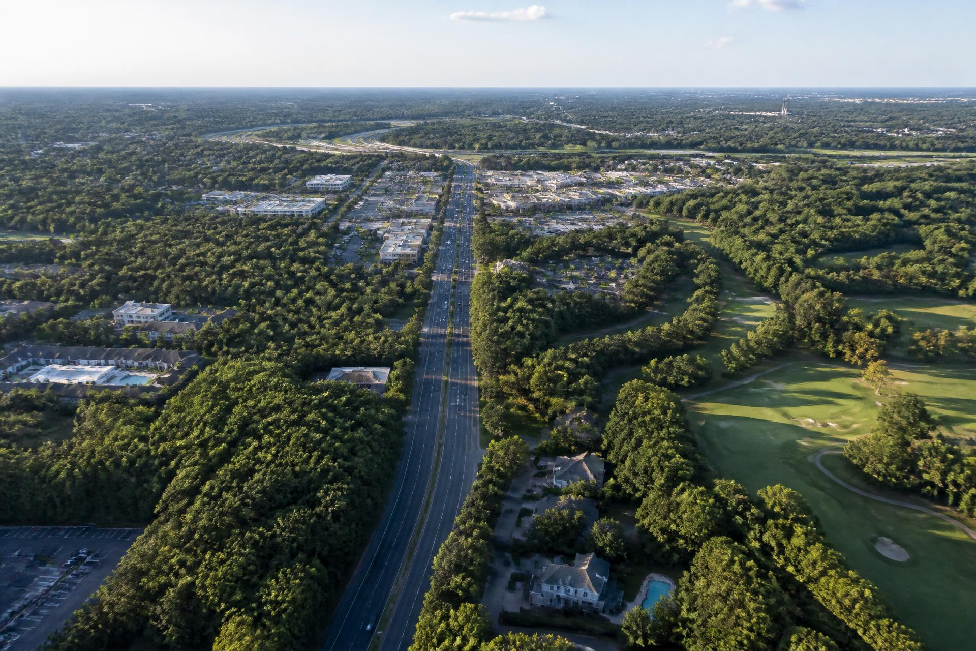 Aerial view of a South Charlotte corridor with a tree-lined arterial road, golf fairways, retail, and the interstate loop on the horizon