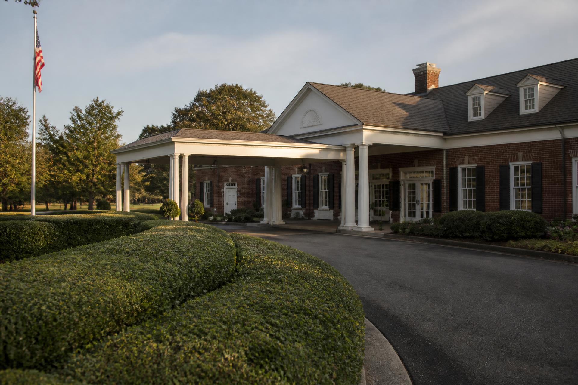 The covered arrival entrance of a brick country club clubhouse with clipped hedges in late-afternoon light
